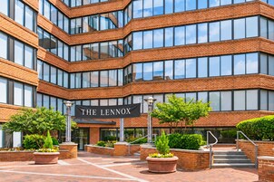 A multi-story building with a brick facade and large windows. There is a sign that reads 'THE LENNOX' and a well-maintained outdoor area with potted plants and a staircase.