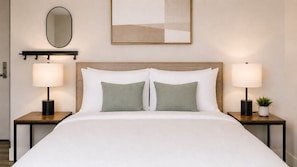 Une chambre d’hôtel avec un grand lit, deux tables de chevet avec des lampes, un miroir et un tableau abstrait encadré accroché au mur.