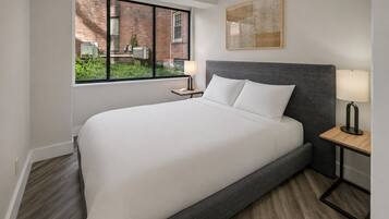 Una habitación de hotel con una cama grande, una mesita de noche con una lámpara, una ventana con vista a un edificio de ladrillo y un cuadro en la pared.