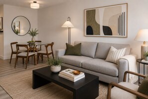 A modern living room with a grey sofa, wooden chairs, a coffee table, a floor lamp, and a painting on the wall.