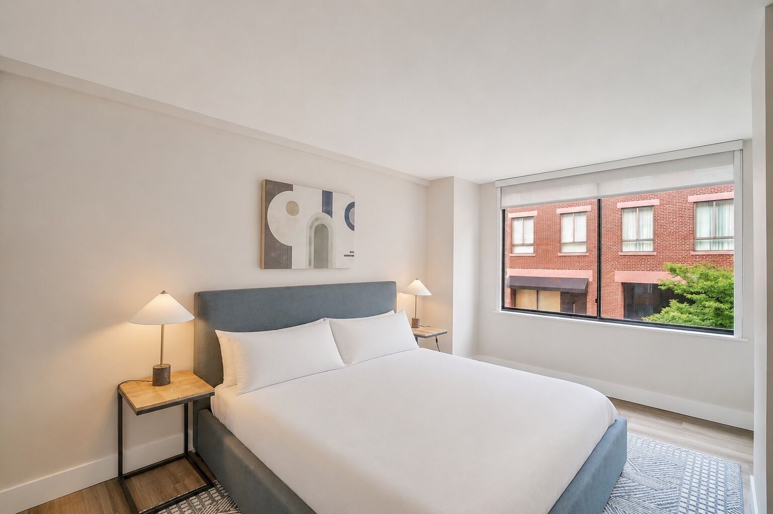 Ein Hotelzimmer mit einem Bett, zwei Nachttischlampen, einem Fenster mit Blick auf Gebäude und einem an der Wand befestigten abstrakten Kunstwerk.