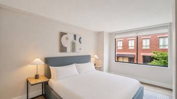Ein Hotelzimmer mit einem Bett, zwei Nachttischlampen, einem Fenster mit Blick auf Gebäude und einem an der Wand befestigten abstrakten Kunstwerk.