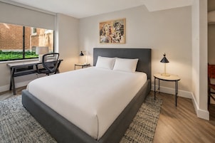 A hotel room with a large bed, a desk, two chairs, a window with a view of greenery, and a painting on the wall.