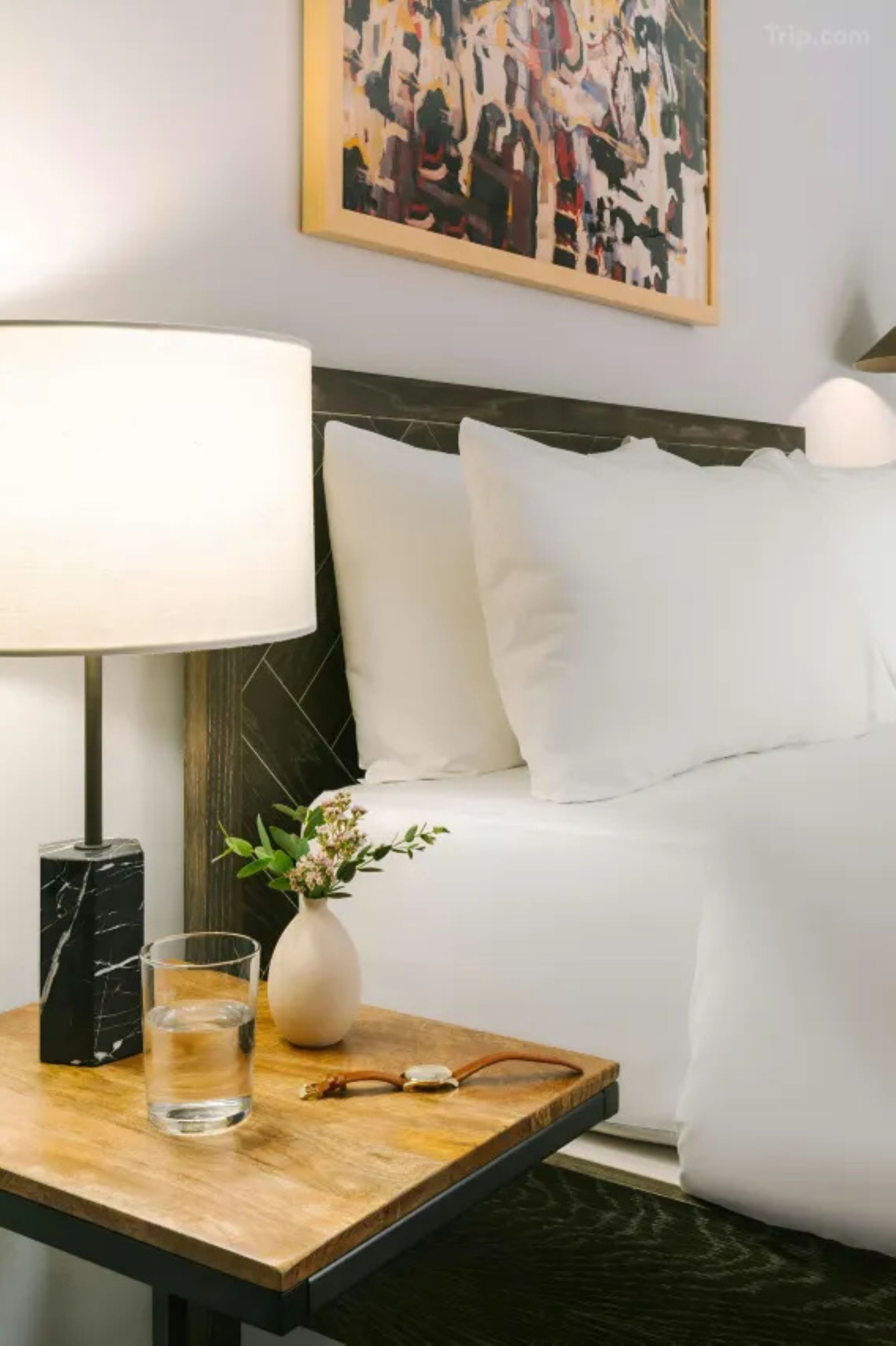 A neatly made bed with white linens, a bedside table with a lamp, a vase with flowers, and a glass of water.