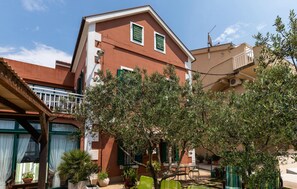 Ein Gebäude mit roter Fassade, grünen Fensterläden und einem Balkon. Blumentöpfe und Gartenmöbel sind sichtbar.