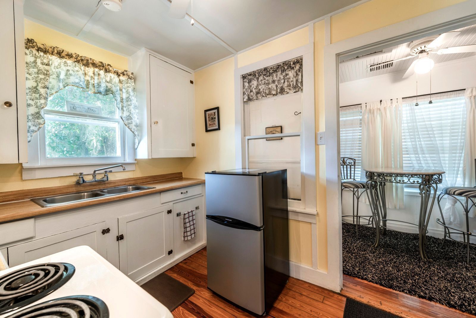 Una cocina compacta con armarios blancos, un refrigerador de acero inoxidable, una encimera de madera y una ventana con cortinas de flores.