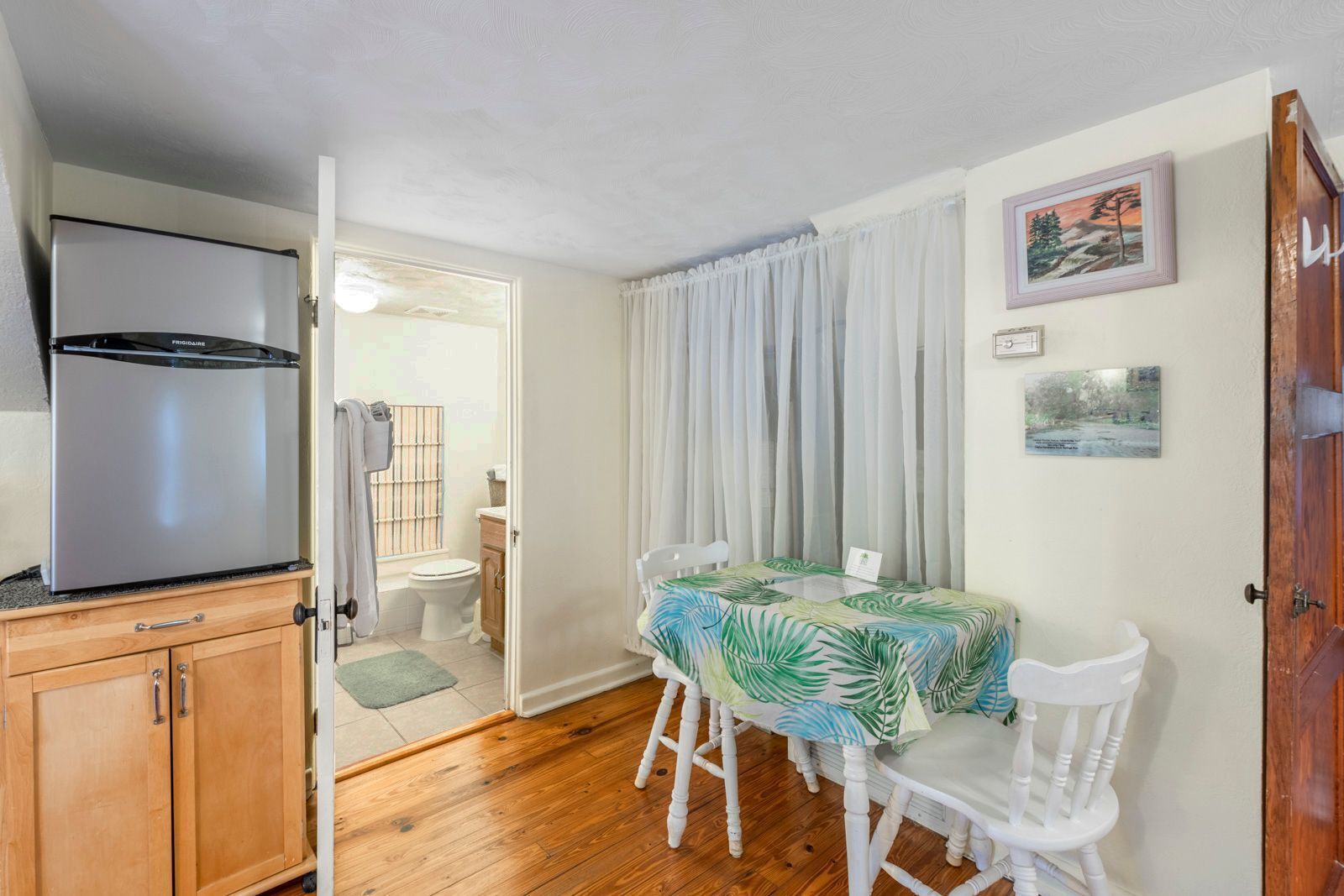 A compact kitchen with a refrigerator, wooden cabinets, a dining table with chairs, and a window with curtains.