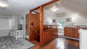A spacious room with a wooden staircase, a kitchen area with wooden cabinets, a small seating area with a chair and a table, and a patterned rug on the floor. - Historic Tremain  Cottages (Mount Dora)