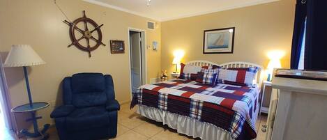A bedroom with a bed, a chair, a ceiling fan, a wall clock, and a lamp.