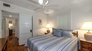 A bedroom with a bed, two bedside tables with lamps, a ceiling fan, and a door leading to a hallway. - Historic Tremain  Cottages (Mount Dora)