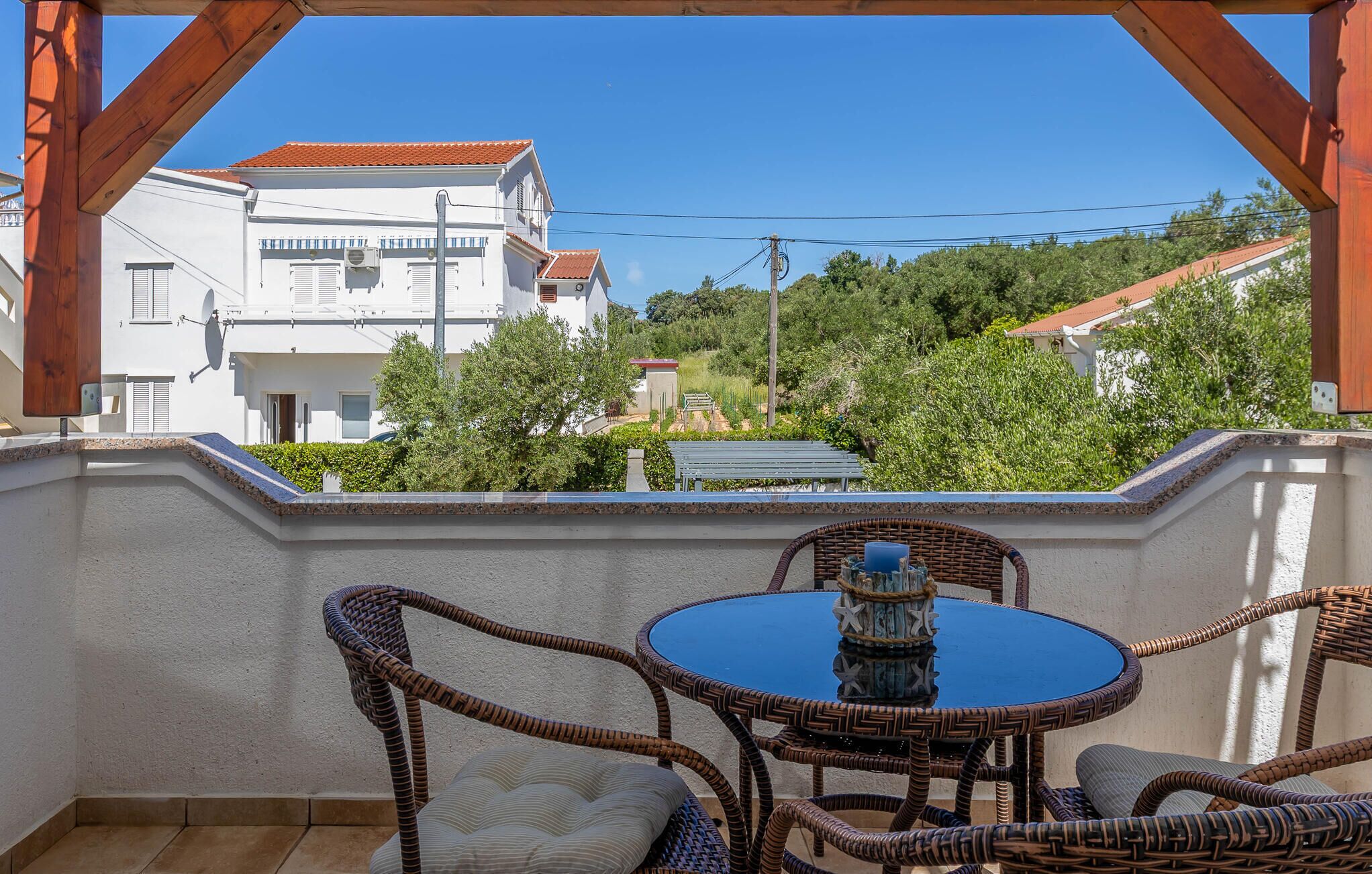 Un balcon doté de meubles en osier et d’une table bleue, donnant sur un quartier résidentiel avec des maisons et de la verdure.