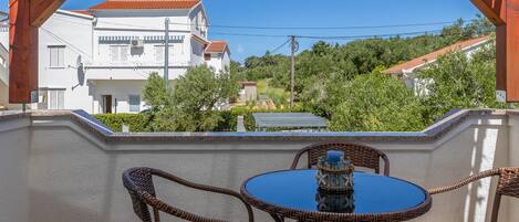 Un balcon doté de meubles en osier et d’une table bleue, donnant sur un quartier résidentiel avec des maisons et de la verdure.