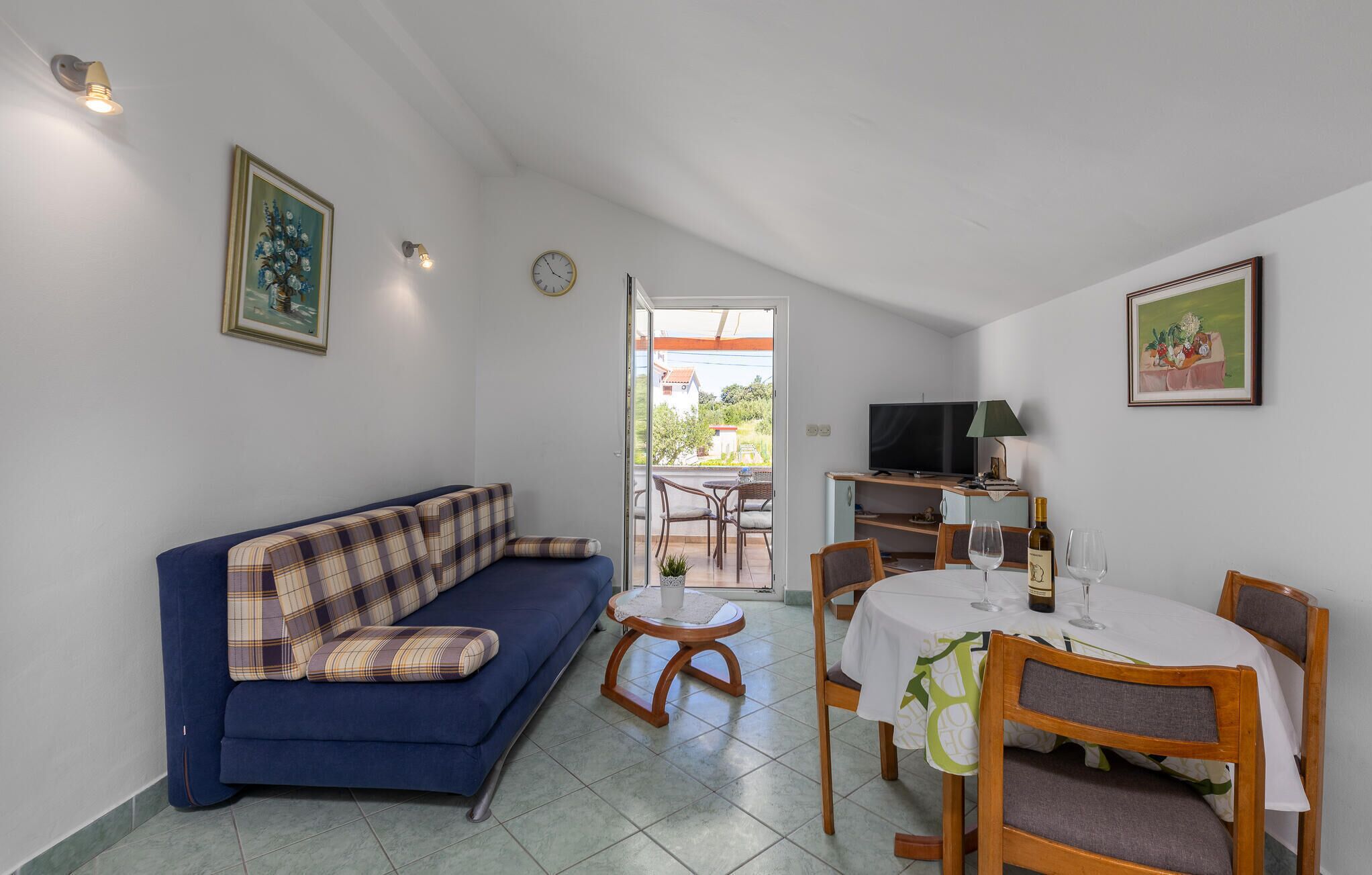 A living room with a blue sofa, a dining table with chairs, a television, and a balcony with a view of the outside.