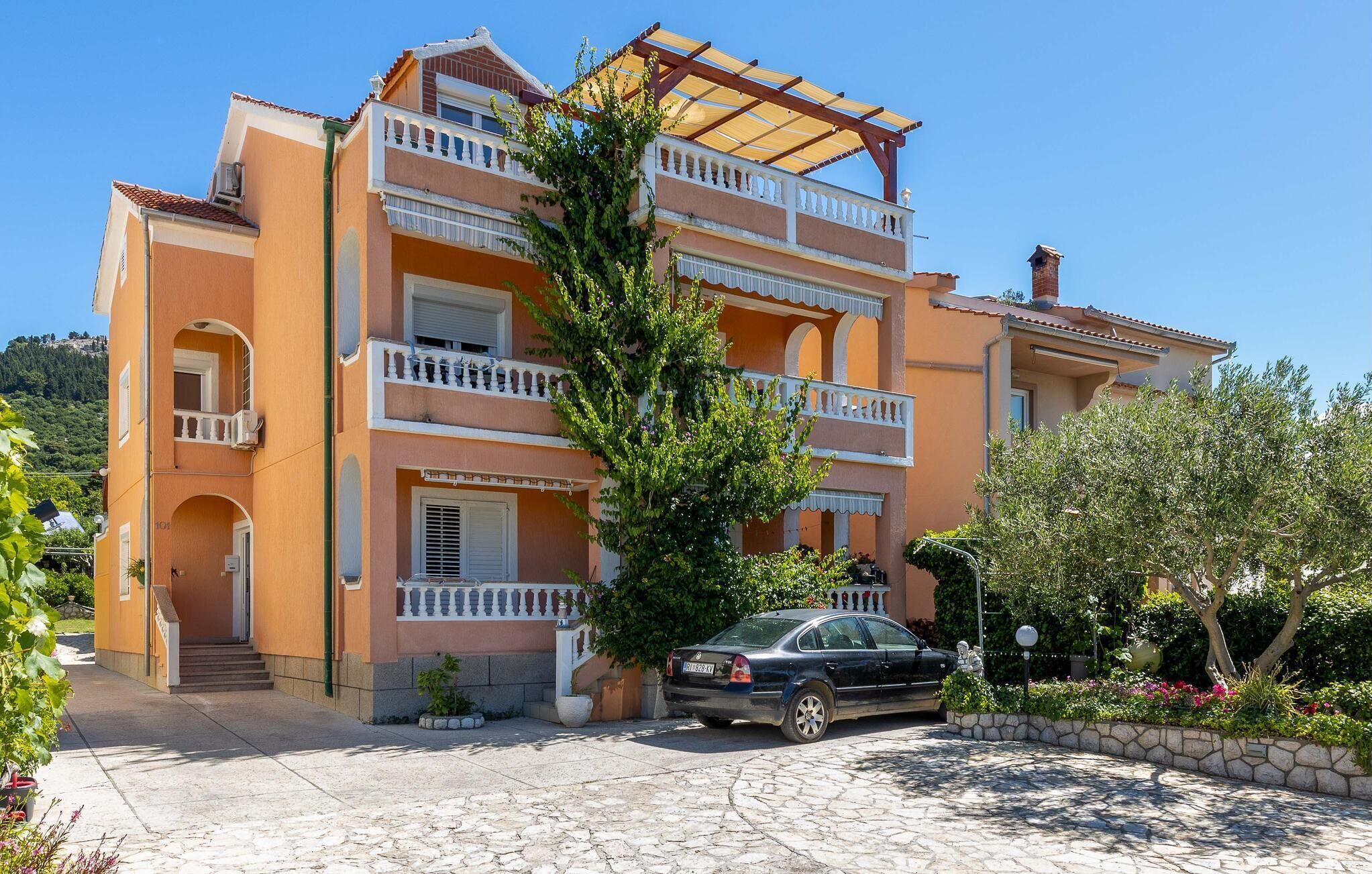 A multi-story building with balconies, a car parked in front, and a stone-paved entrance.