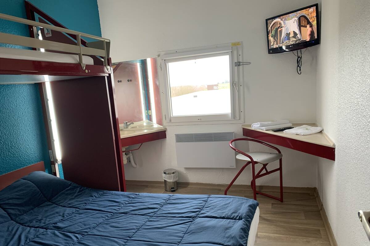Een kleine kamer met een stapelbed, een bureau met stoel, een raam en een wandtelevisie.