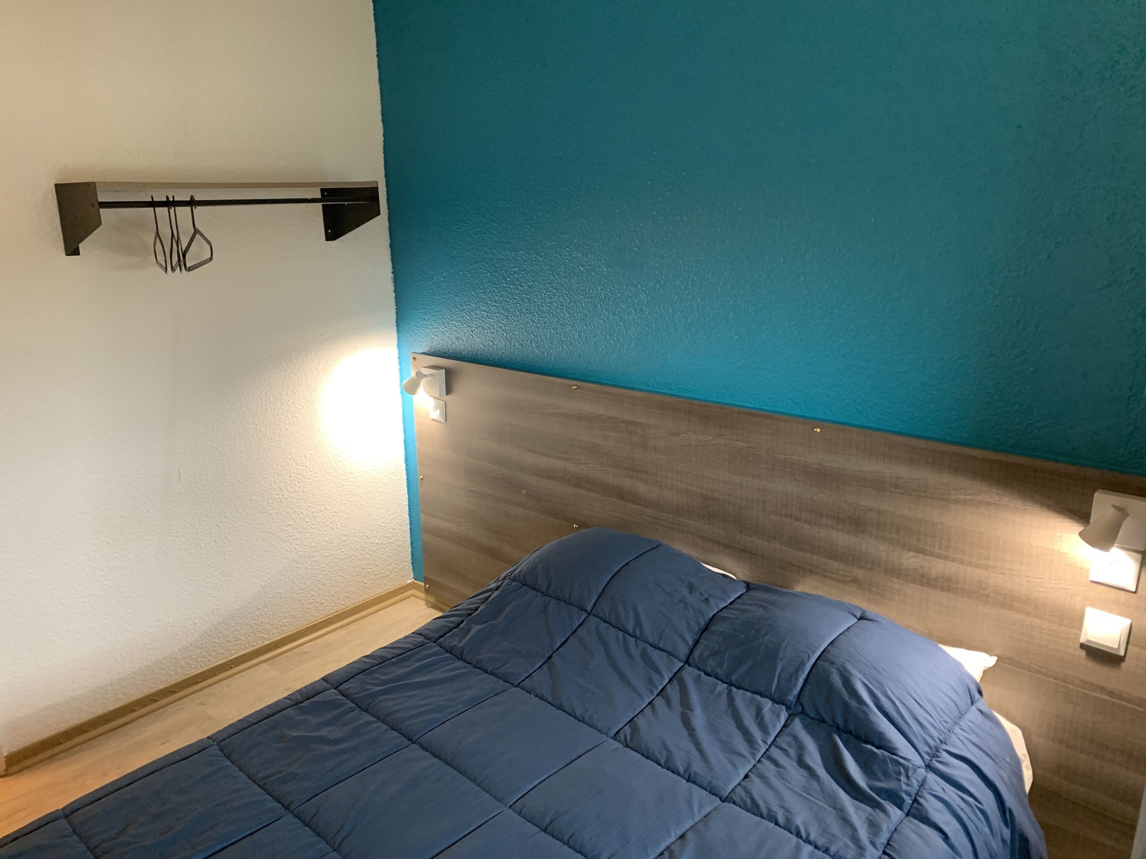 Un dormitorio con cama, cabecera de madera, lámpara de pared y estante con ganchos.