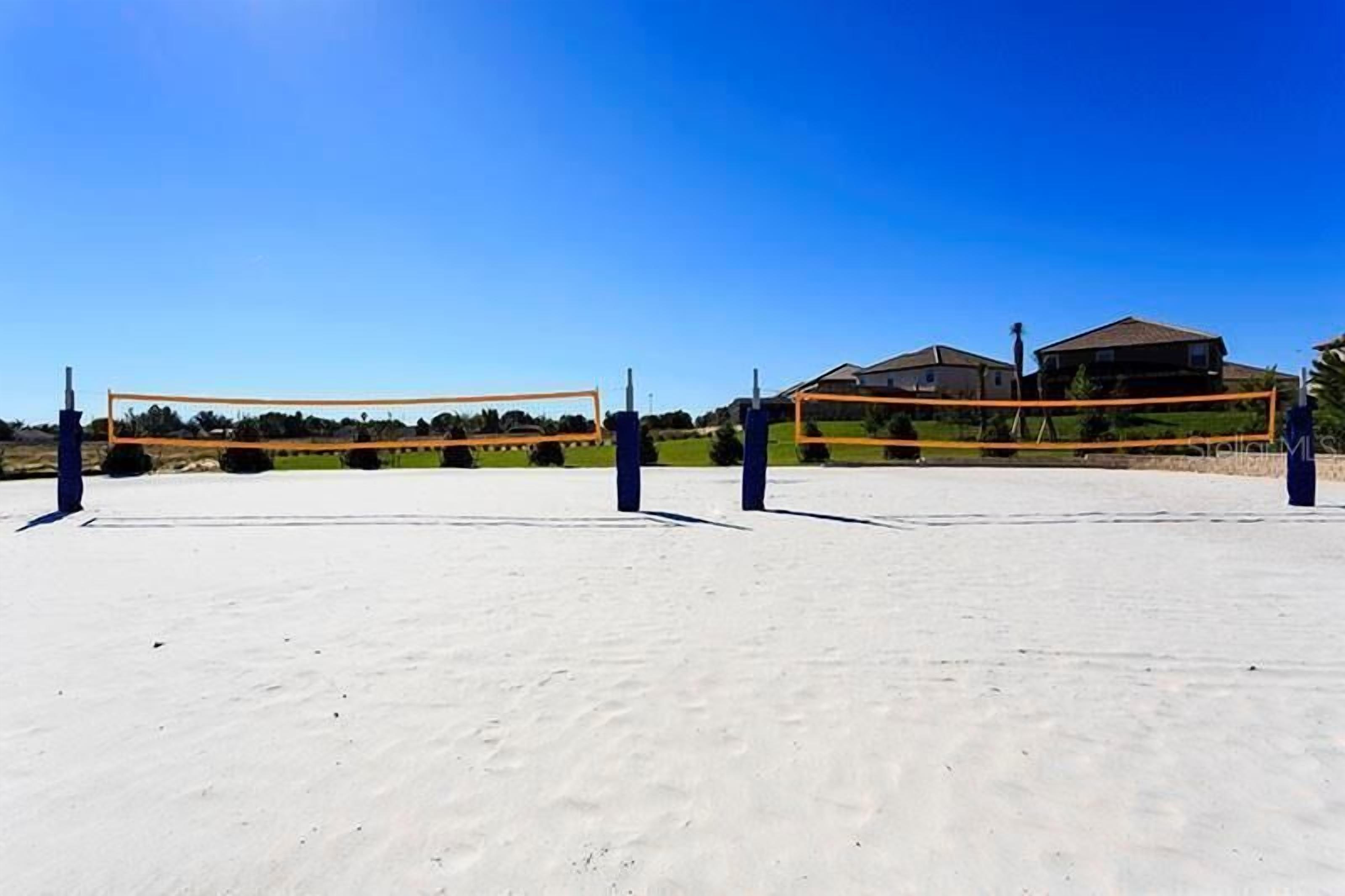 A volleyball court with a net, surrounded by a sandy area and houses in the background.