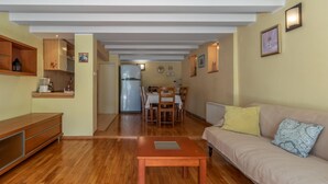 Ein Wohnzimmer mit Holzboden, einer Couch mit gemusterten Kissen, einem Couchtisch und einer Küchenzeile mit Kühlschrank und Esstisch.