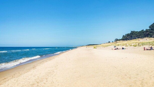 Ein weiter Sandstrand mit sanften Wellen, blauem Himmel und einigen entspannten Personen.