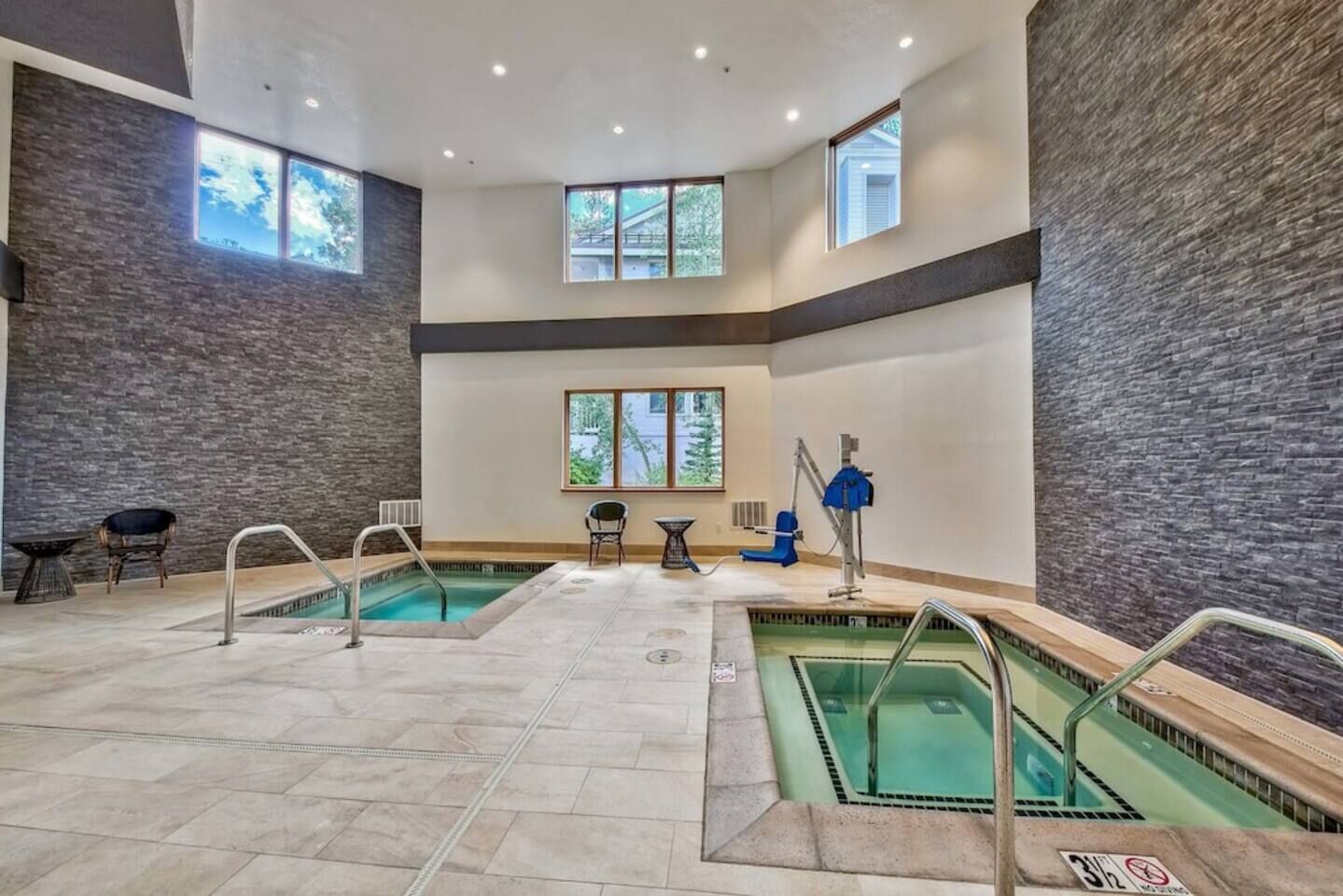 Ein moderner Poolbereich im Innenbereich mit zwei Pools, Sitzgelegenheiten und Blick auf Bäume durch Fenster.