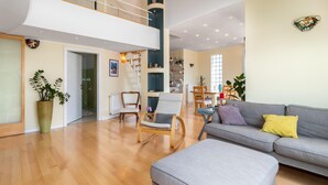 A spacious living room with a grey sofa, a wooden coffee table, and a grey ottoman. There are two rocking chairs and a dining table with chairs. A large window provides natural light, and a staircase leads to an upper level. - Lovely apartment in Crikvenica (Crikvenica)