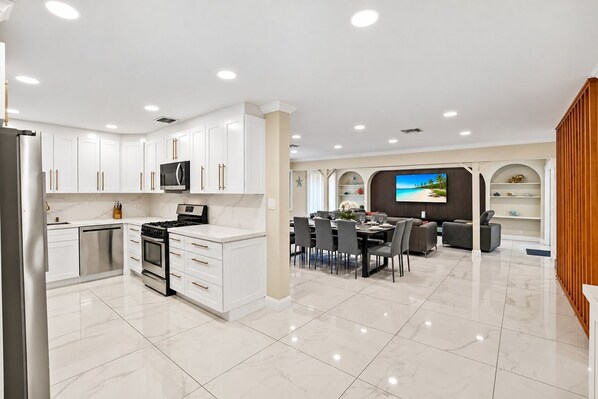 A modern kitchen with white cabinetry, a built-in microwave, and a dining area with a large TV displaying a beach scene.