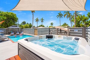 A hot tub and a pool with lounge chairs and umbrellas, surrounded by palm trees and a wooden fence.