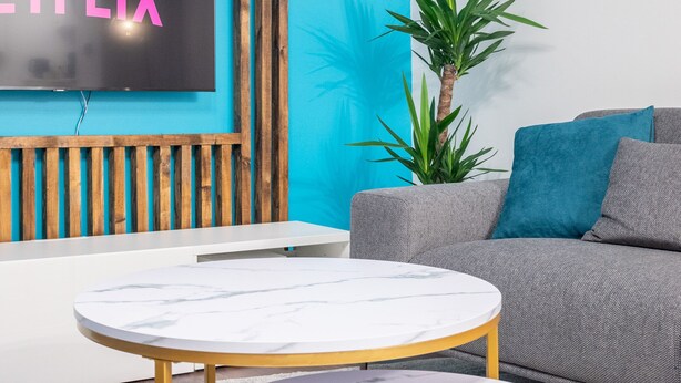 A modern living room with a large flat-screen TV displaying "NETFLIX," a grey sofa with blue cushions, a small potted plant, and two round marble tables with gold legs. - Le Moja Tourcoing (Tourcoing)