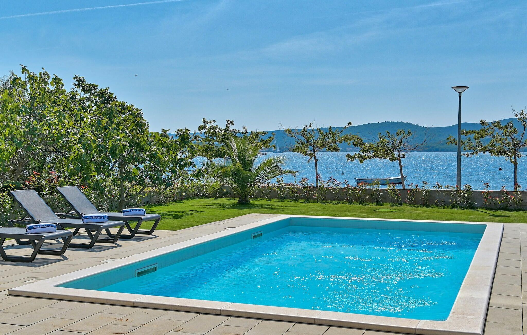 En rektangulär pool med ett klart, blått vatten, omgiven av en kakelbeklädd terrass. Här finns solstolar med handdukar märkta med texten "Villa Roberta" och en lyktstolpe. I bakgrunden syns en vattenyta med en båt och ett bergskedja i fjärran.