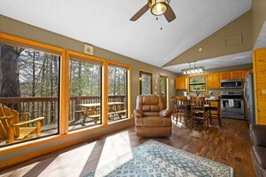A spacious living room with large windows, a ceiling fan, a fireplace, and a kitchen area with wooden cabinets and appliances.