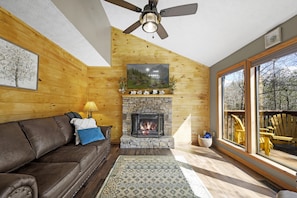 A cozy living room with a stone fireplace, a ceiling fan, and a large window offering a view of the outdoors.