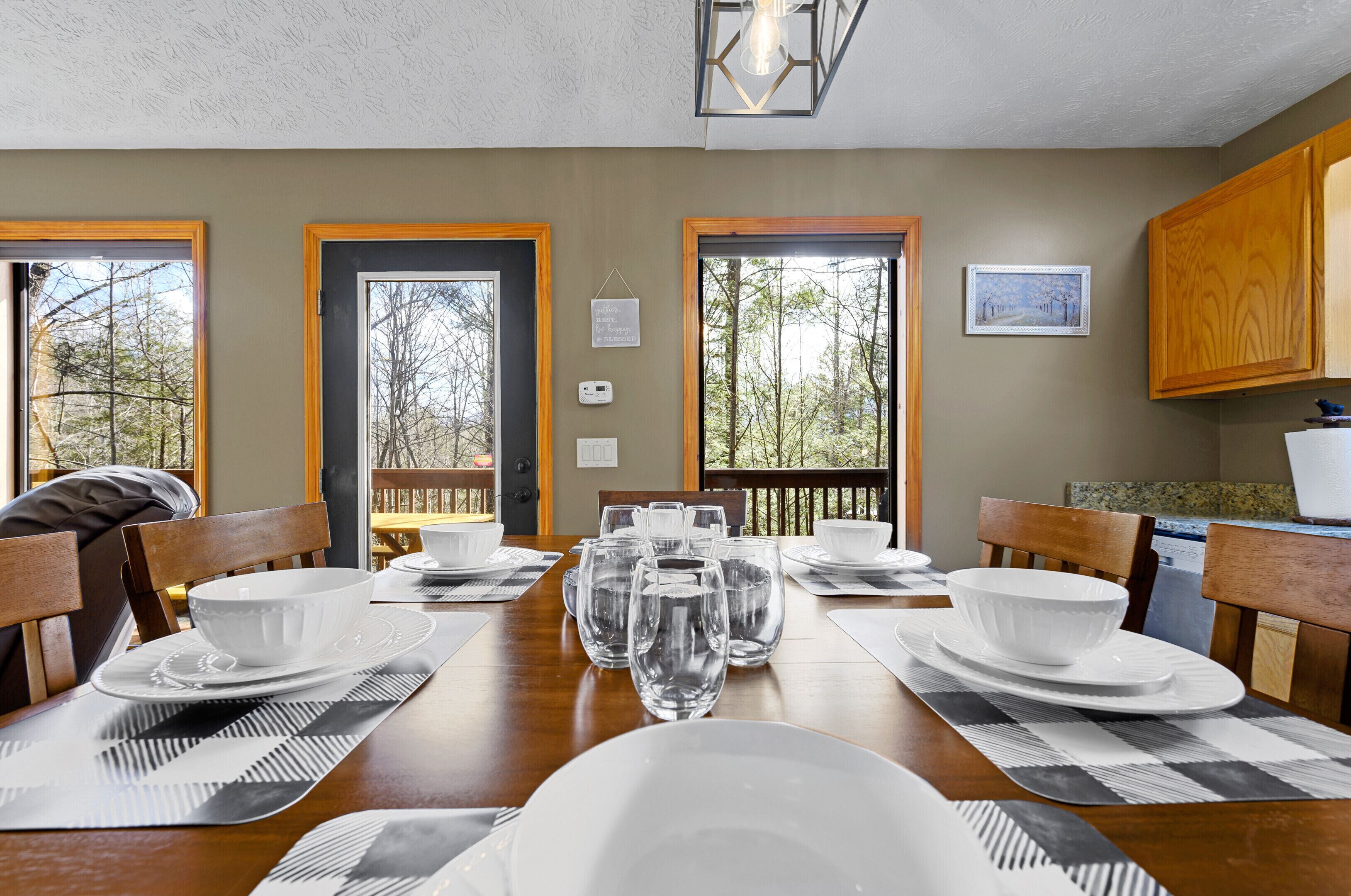 Un comedor con una mesa de madera dispuesta para cuatro personas, rodeada de sillas. Hay varias copas de vino y cuencos sobre la mesa. La habitación tiene ventanas amplias con vistas a árboles y una terraza exterior.