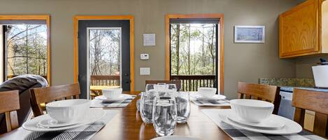 Un comedor con una mesa de madera dispuesta para cuatro personas, rodeada de sillas. Hay varias copas de vino y cuencos sobre la mesa. La habitación tiene ventanas amplias con vistas a árboles y una terraza exterior.