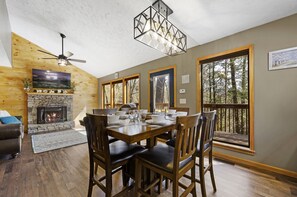 L’intérieur chaleureux d’un chalet avec une cheminée en pierre, une table à manger en bois et de grandes fenêtres donnant sur la forêt.