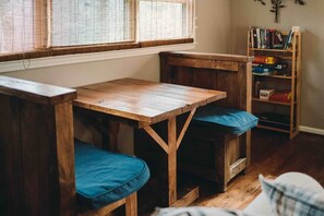 Un banco de madera con asiento acolchado y una mesita en una habitación con una estantería y una ventana con persianas.