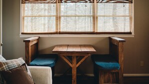 Una habitación acogedora con una mesa de madera y dos sillas, un sofá con cojines estampados y una ventana con persianas.