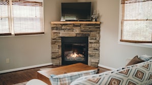 Un salón con chimenea de piedra, un televisor de pantalla plana encima, una mesa de centro de madera y un plaid estampado sobre un sofá blanco.