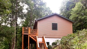 Una cabaña de troncos con una escalera de madera que lleva a una terraza, rodeada de un bosque frondoso.