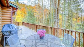 Una terraza de madera con una mesa y sillas de metal, rodeada por un pasamanos de madera, con vistas a un bosque de árboles con colores otoñales.