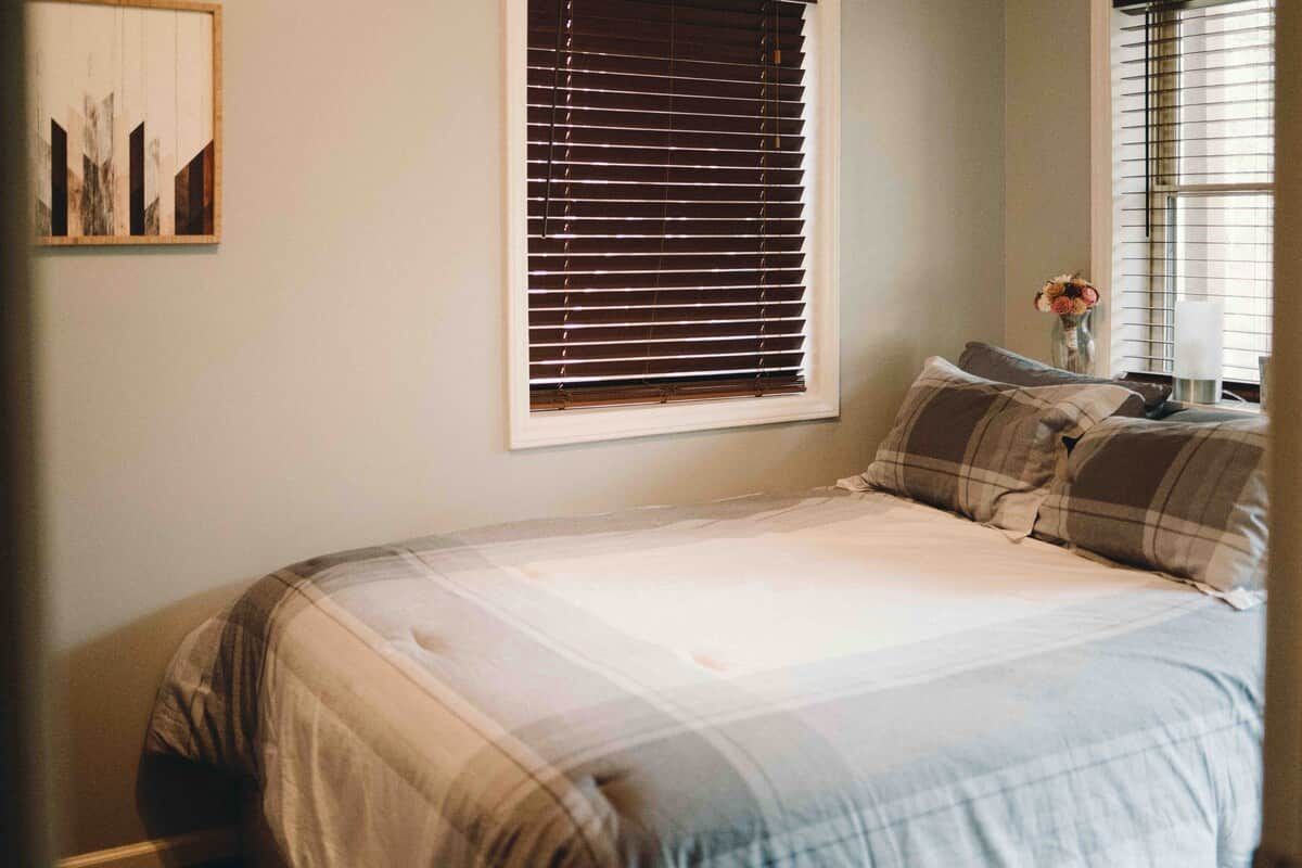 Une chambre à coucher avec un lit, une fenêtre avec des stores, une œuvre d’art accrochée au mur et un vase rempli de fleurs.