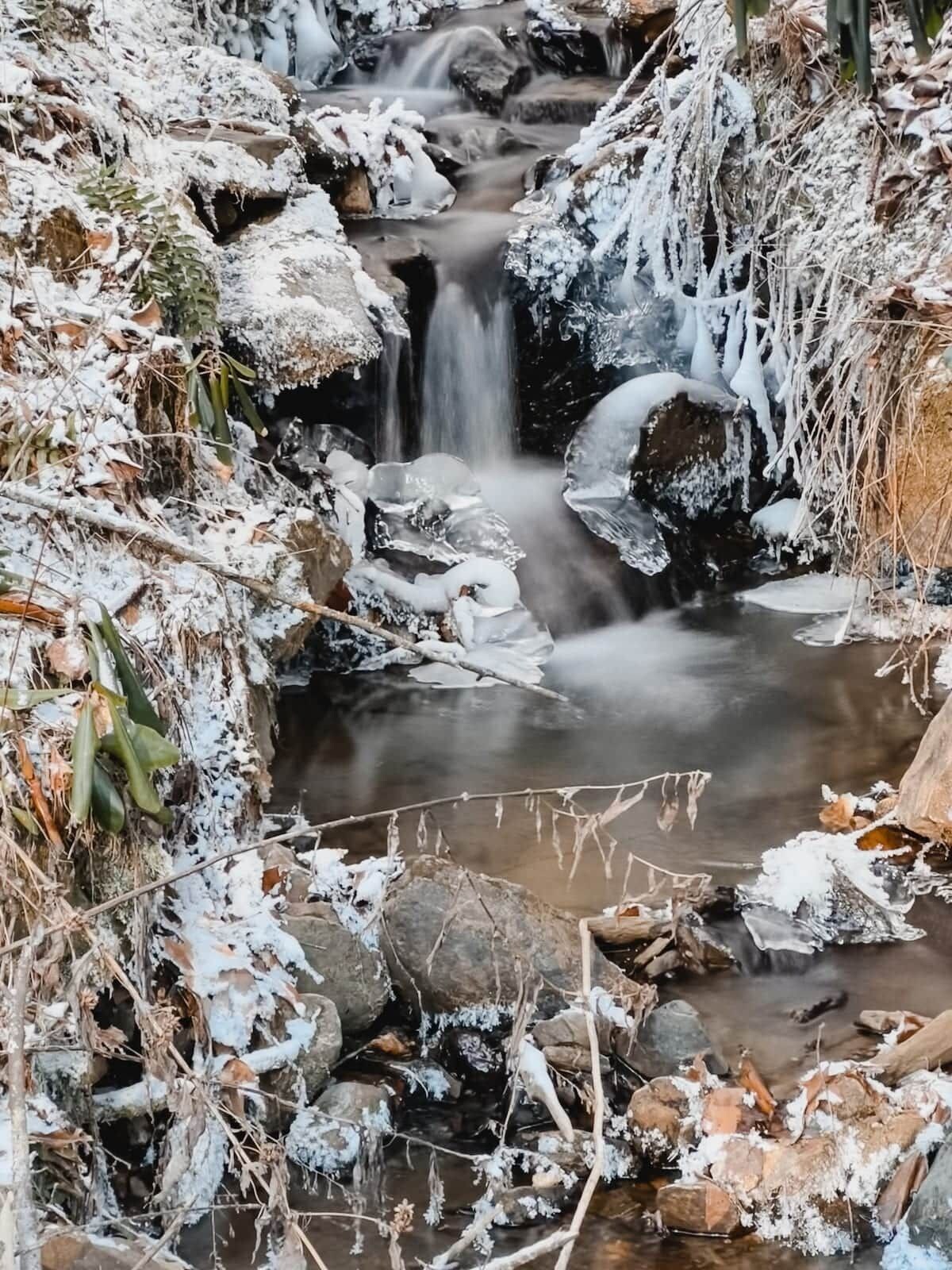 A natural landscape with a waterfall, rocks, and snow-covered vegetation.