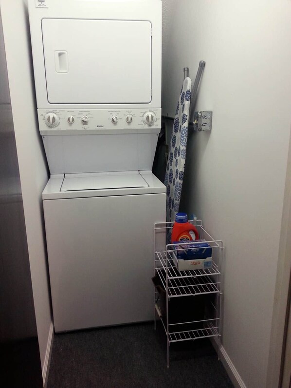 A compact laundry room with a front-loading washing machine, a drying rack, and a metal shelf holding cleaning supplies.