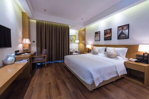 Habitación de hotel con una cama grande, mesitas de noche, un escritorio y una silla. Hay cortinas, cuadros en la pared y un televisor montado.
