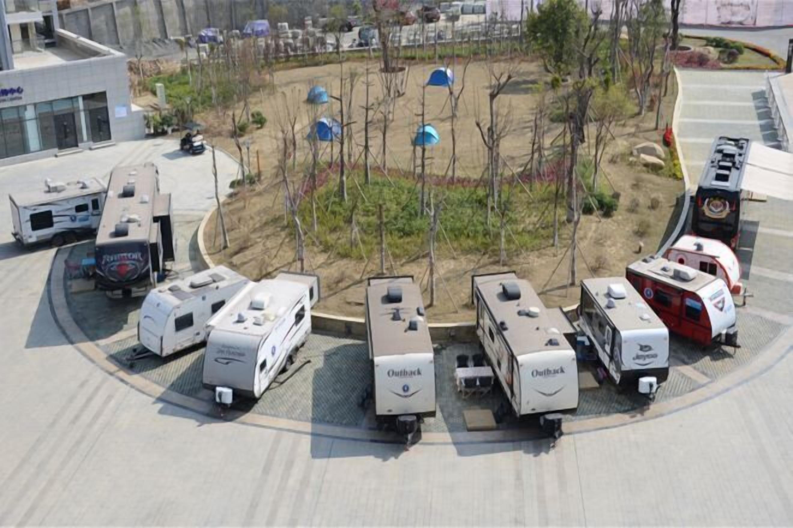 Un espace de stationnement où sont garés plusieurs camping-cars et remorques, un jardin aménagé avec des arbres et des arbustes, et un bâtiment moderne en arrière-plan.