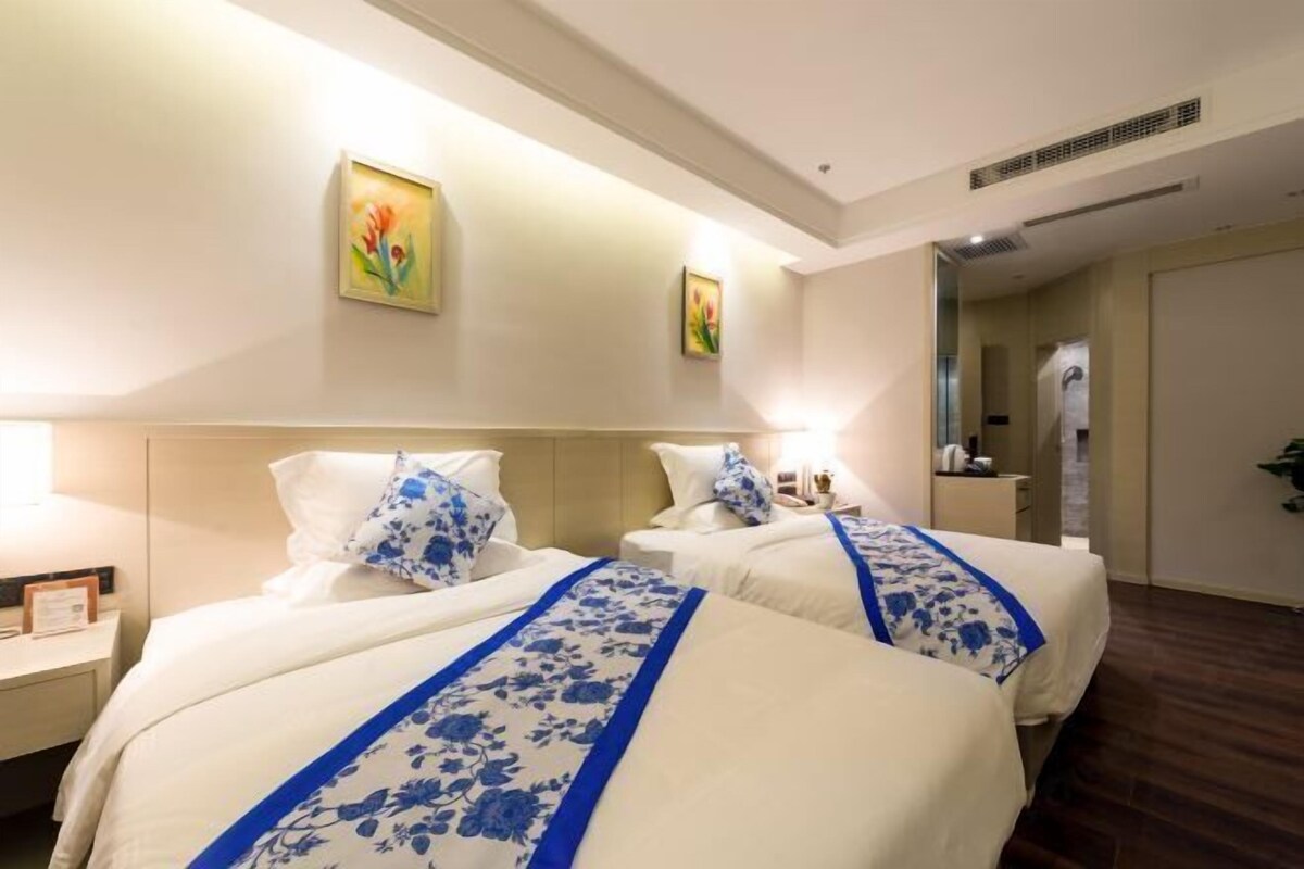 Habitación de hotel con dos camas, cada una con ropa blanca de cama y detalles florales azules, una mesita de noche con lámpara y un cuadro en la pared.