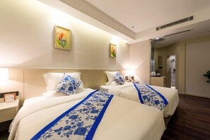 Habitación de hotel con dos camas, cada una con ropa blanca de cama y detalles florales azules, una mesita de noche con lámpara y un cuadro en la pared.