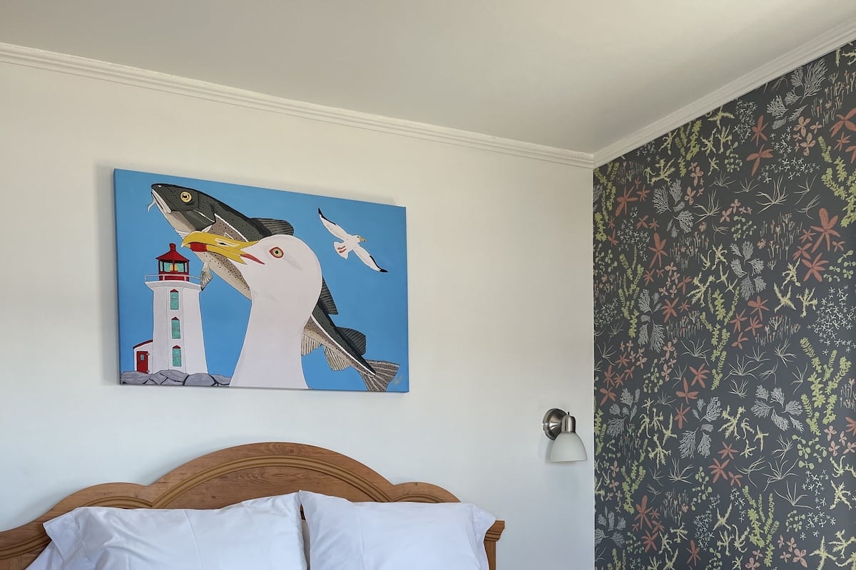 Ein ordentlich bezogenes Bett mit weißen Kissen und einer grauen, strukturierten Tagesdecke, ein hölzernes Kopfteil, ein Wandbild von einem Möwen und Leuchtturm sowie eine Wandtapete mit Muster.