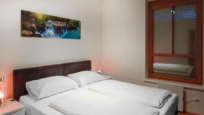 Una habitación de hotel con cama, mesita de noche, un cuadro colgado en la pared y una ventana con persianas.