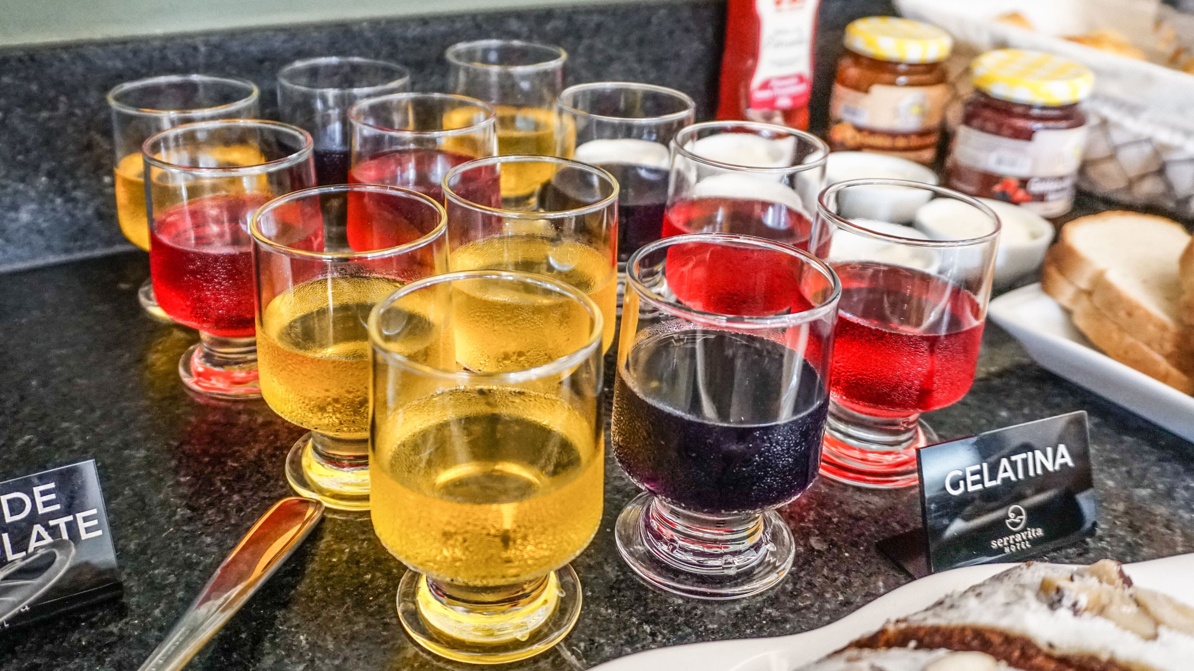 A selection of colorful beverages in glasses on a counter, with a jar of jelly and a plate of bread.