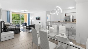 A modern living room with a glass dining table, white chairs, a sofa, and a balcony with blue curtains. - Yacht Club Miami Suite 2 Bedroom and 2 Bathroom With Baby Crib & Free Parking (Aventura)